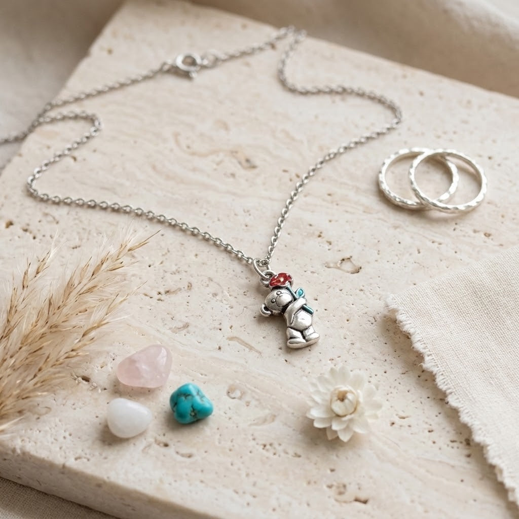 A silver-tone necklace with a small bear charm holding a red and green hat, displayed on a textured beige stone slab with dried grass, crystals, and two textured rings.