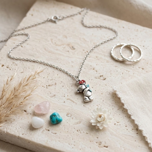 A silver-tone necklace with a small bear charm holding a red and green hat, displayed on a textured beige stone slab with dried grass, crystals, and two textured rings.