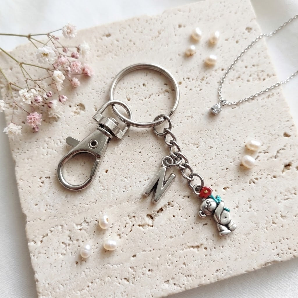A silver-toned keychain with a lobster clasp, a letter charm, and a small teddy bear charm with red and teal accents, displayed on a textured cream stone tile with dried flowers and small pearls.