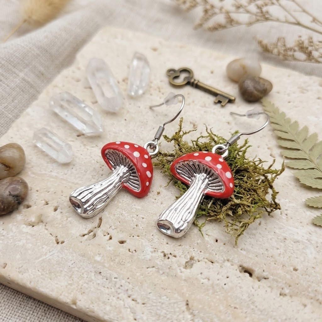 A pair of dangling mushroom earrings with red caps, white spots, and silver stems, displayed on a stone surface with crystals, moss, dried leaves, and a small key.