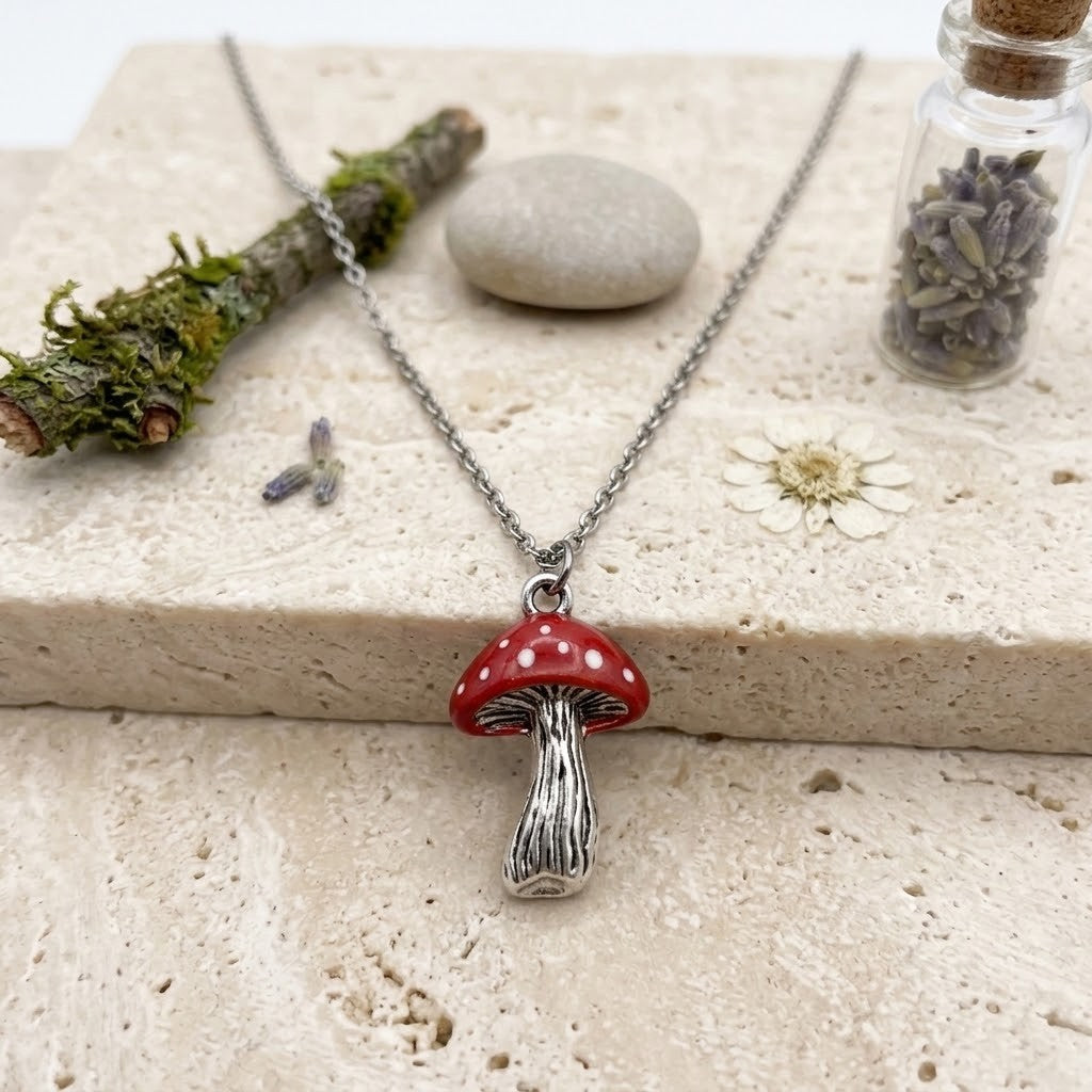 A silver chain necklace with a red and white spotted mushroom pendant, styled on a beige stone slab with a mossy twig, a smooth stone, a small glass vial, and dried lavender.