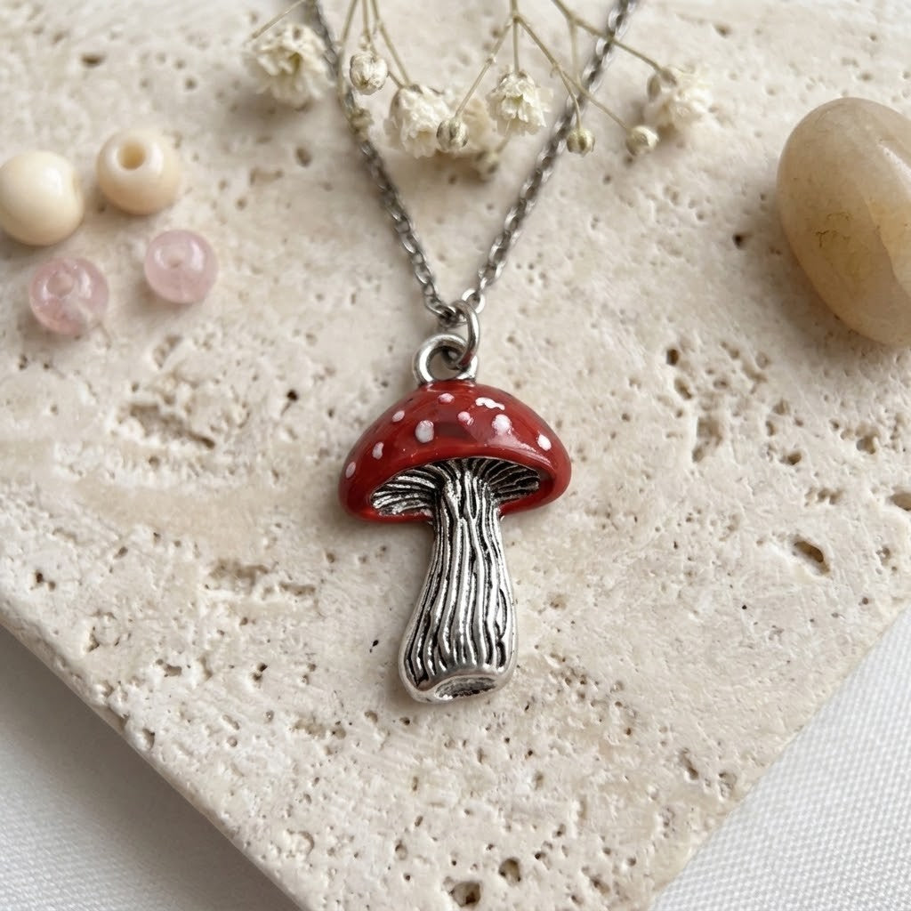 A silver-toned mushroom pendant necklace with a red cap and white spots, displayed on a textured beige stone surface with small dried flowers and beads.
