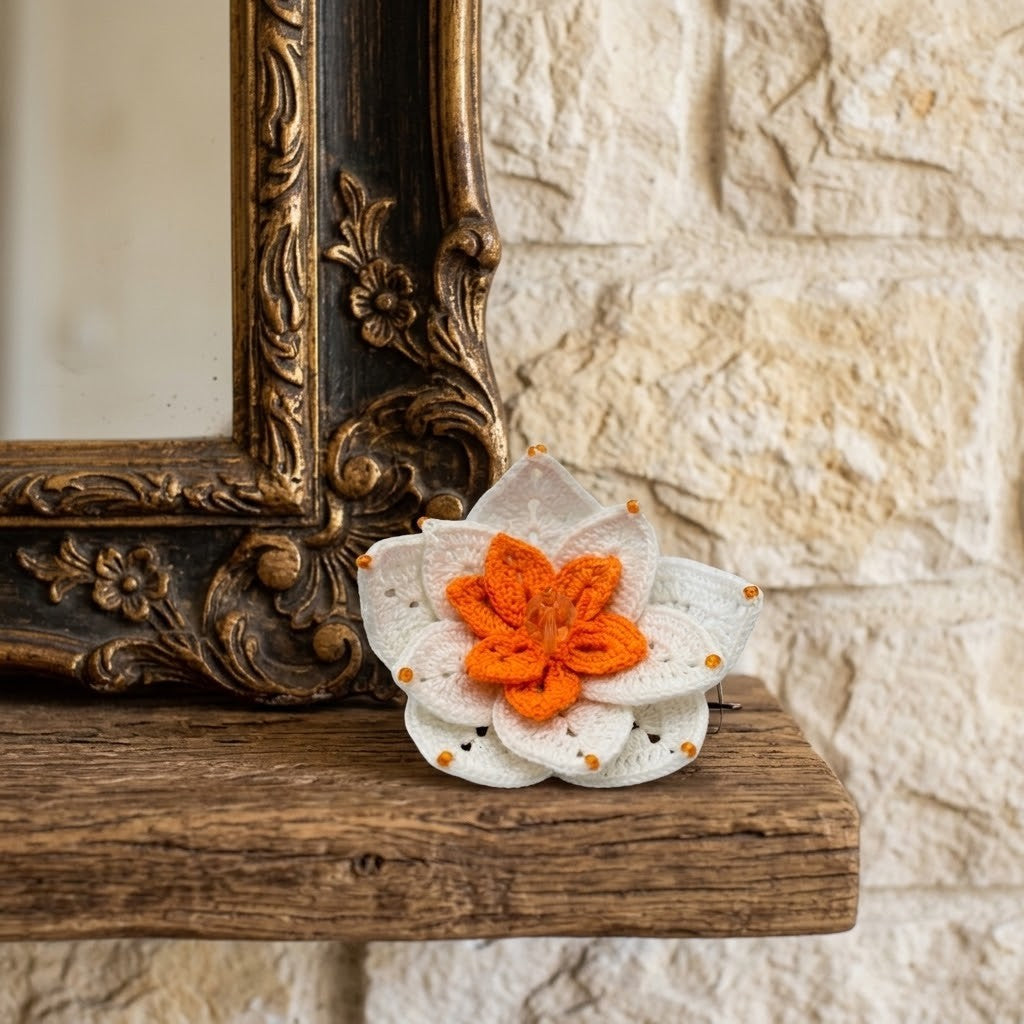 A crocheted waterlily brooch with white petals and an orange centre, resting on a rustic wooden mantel in front of a stone wall and an ornate gold mirror frame.