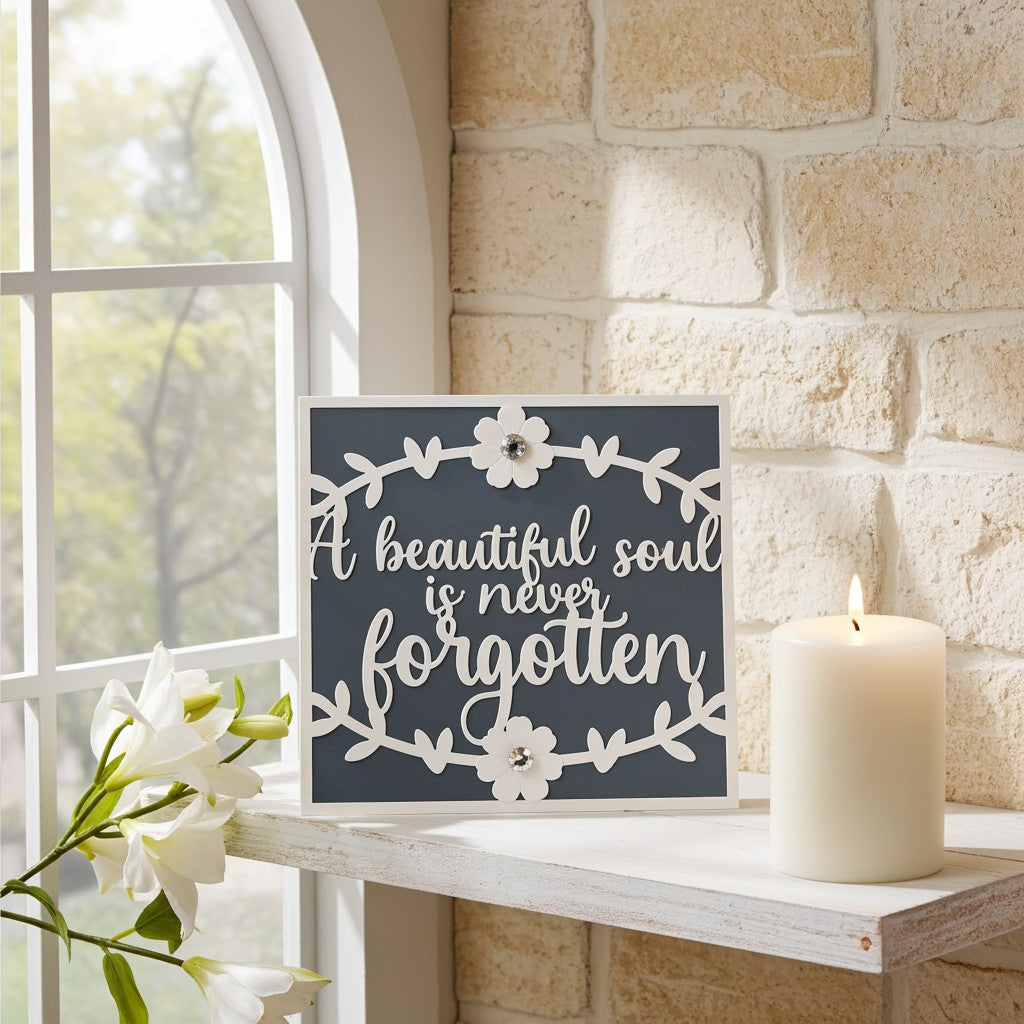 A square decorative sympathy card with the phrase “A beautiful soul is never forgotten” in white script on a dark charcoal background, styled on a white shelf beside a lit cream candle and white flowers against a stone wall and window.