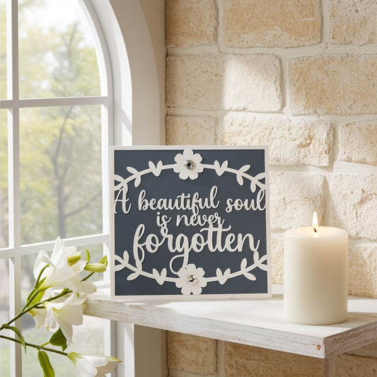 A square decorative sympathy card with the phrase “A beautiful soul is never forgotten” in white script on a dark charcoal background, styled on a white shelf beside a lit cream candle and white flowers against a stone wall and window.