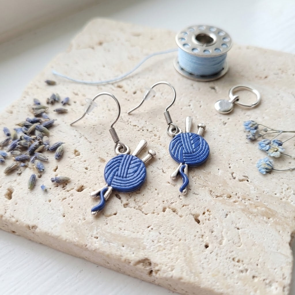A pair of silver-toned dangle earrings shaped like blue yarn balls with knitting needles, displayed on a textured beige stone slab with lavender buds, a spool of light blue thread, and a small silver charm.