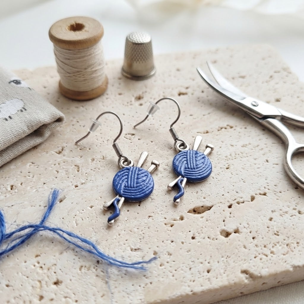 A pair of silver dangle earrings shaped like blue yarn balls with knitting needles, displayed on a textured stone slab with sewing and crafting props including spools of thread, scissors, and fabric.