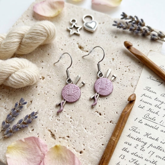 A pair of silver dangle earrings shaped like knitting needles and a ball of lavender yarn, displayed on a textured stone surface with knitting props including a crochet hook, yarn skeins, lavender sprigs, and a printed booklet page.