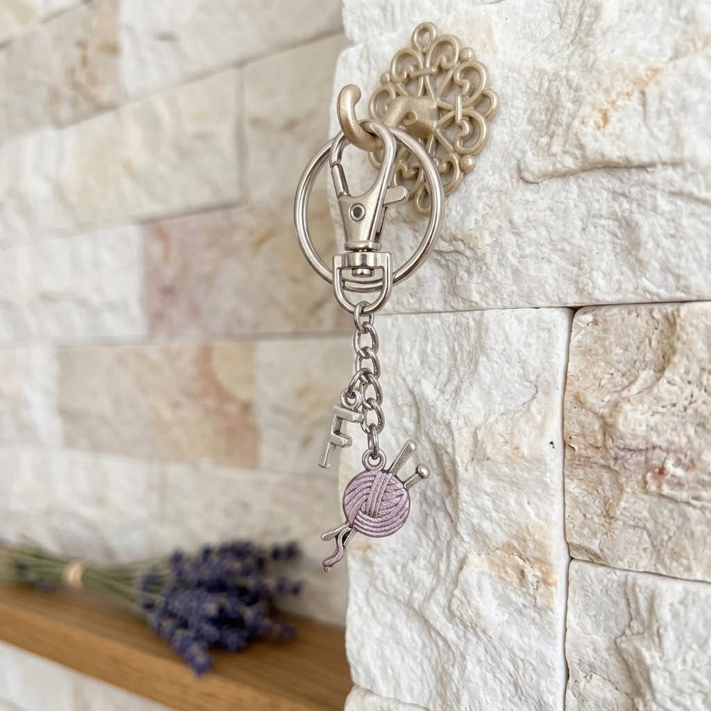 A silver-toned metal keychain with a clasp, ring, and dangling charm shaped like a ball of yarn with knitting needles, shown hanging from a decorative wall hook against a light stone wall.