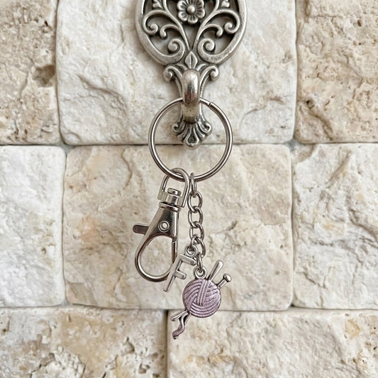 A silver-toned metal keychain with a lobster clasp, key ring, and two dangling charms: a small cross and a lavender-pink ball of yarn with knitting needles. The keychain is displayed hanging from an ornate silver wall hook against a beige stone tile background.