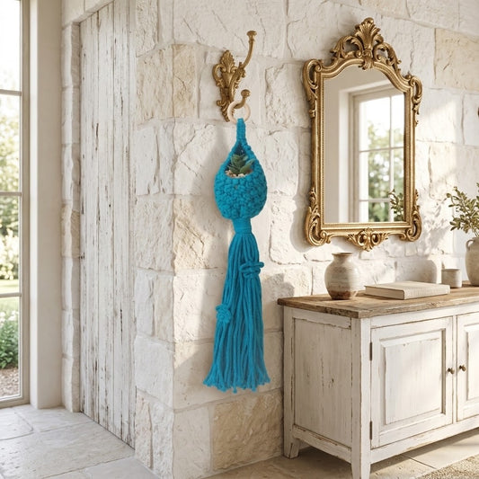 A bright turquoise blue macrame wall hanging with a tassel is displayed on a gold wall hook in a rustic, light stone entryway next to a distressed white cabinet and an ornate gold mirror.
