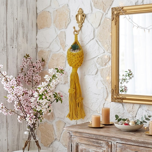 A yellow macrame wall hanging with a tassel and small planter, displayed on a rustic stone wall above a wooden console table with candles, flowers, and a mirror.