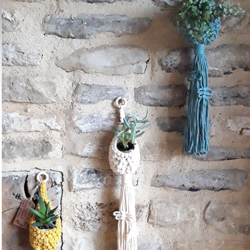 A hanging plant holder with a macrame design, in a shade of blue, displayed on a stone wall with other decorative items.