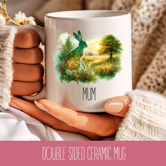 A ceramic coffee mug with a double-sided design featuring a green hare with a rabbit hole in the background, and the word 'MUM' at the bottom.