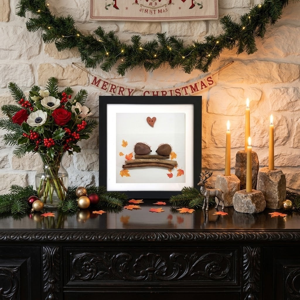 A framed hedgehog-themed pebble art frame featuring a heart, a small red heart, and a couple of hedgehogs with autumn leaves, displayed on a dark wooden mantel in a decorated holiday setting with candles, a flower arrangement, and a stone wall background.