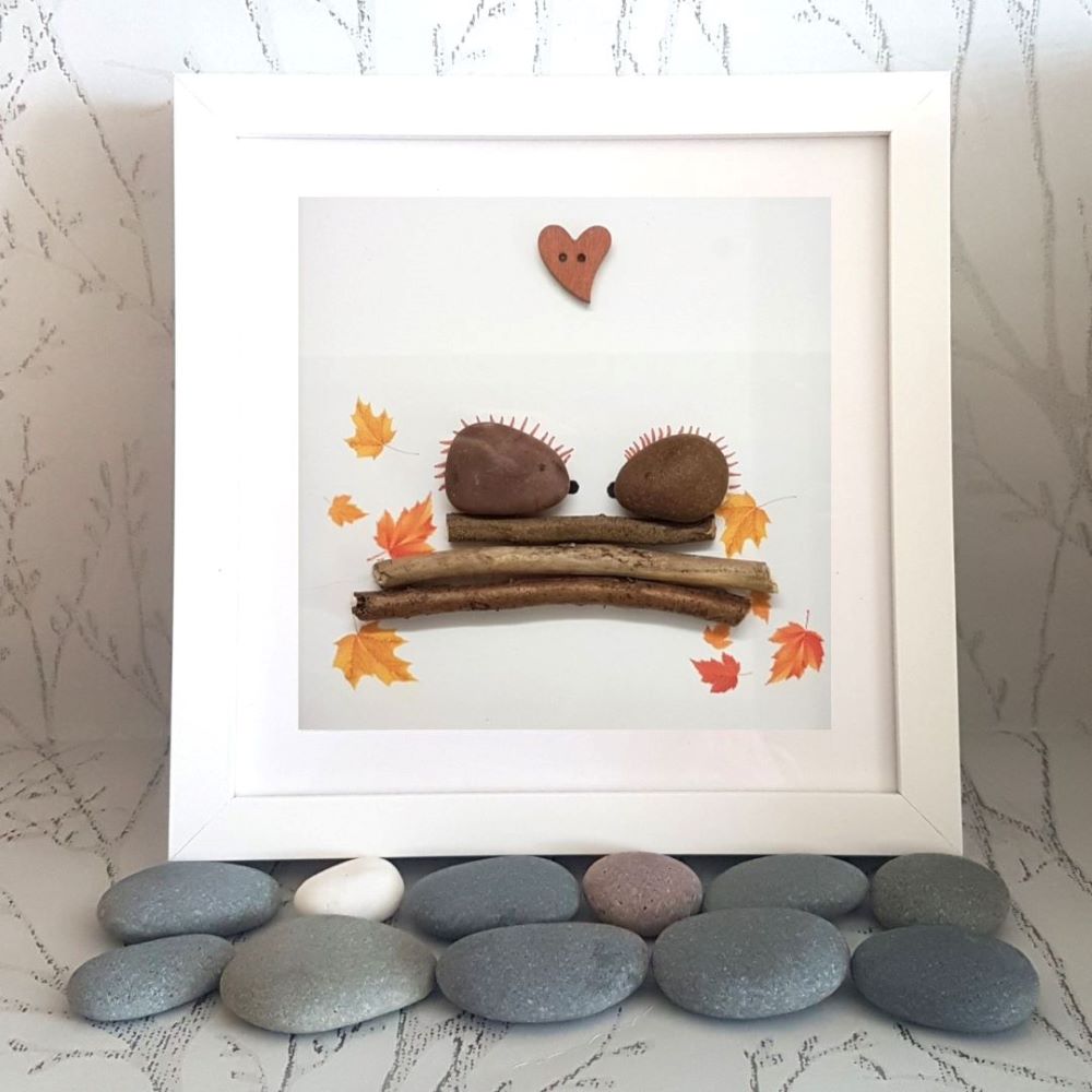 A decorative pebble art picture frame containing a pebble and two silk leaves in the shape of a hedgehog and acorns, set against a white background, with a heart-shaped decoration at the top.