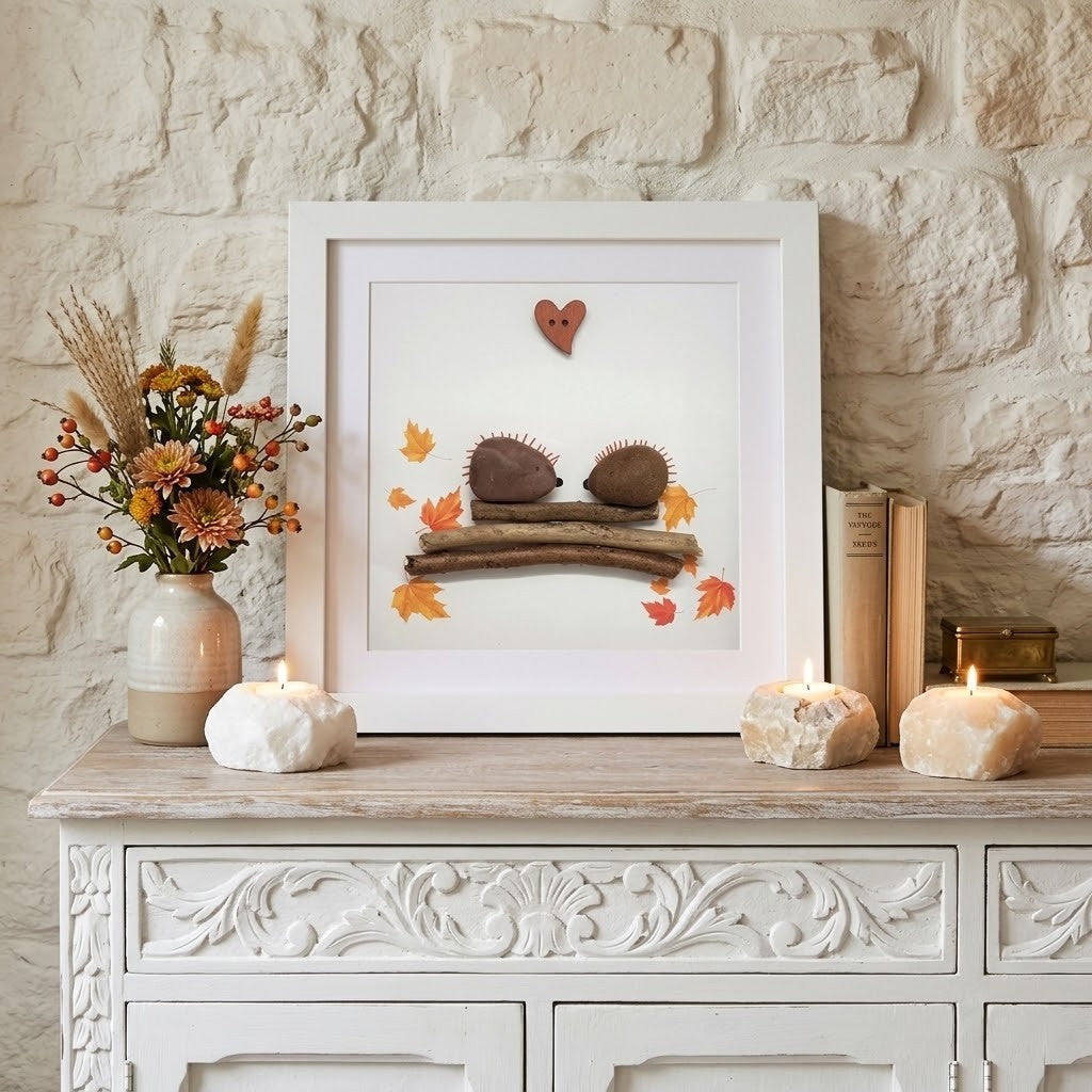 A framed autumn-themed art print displayed on a white carved wooden cabinet against a light stone wall. The print features two small brown hedgehogs on a piece of wood with scattered orange and yellow leaves and a small heart.