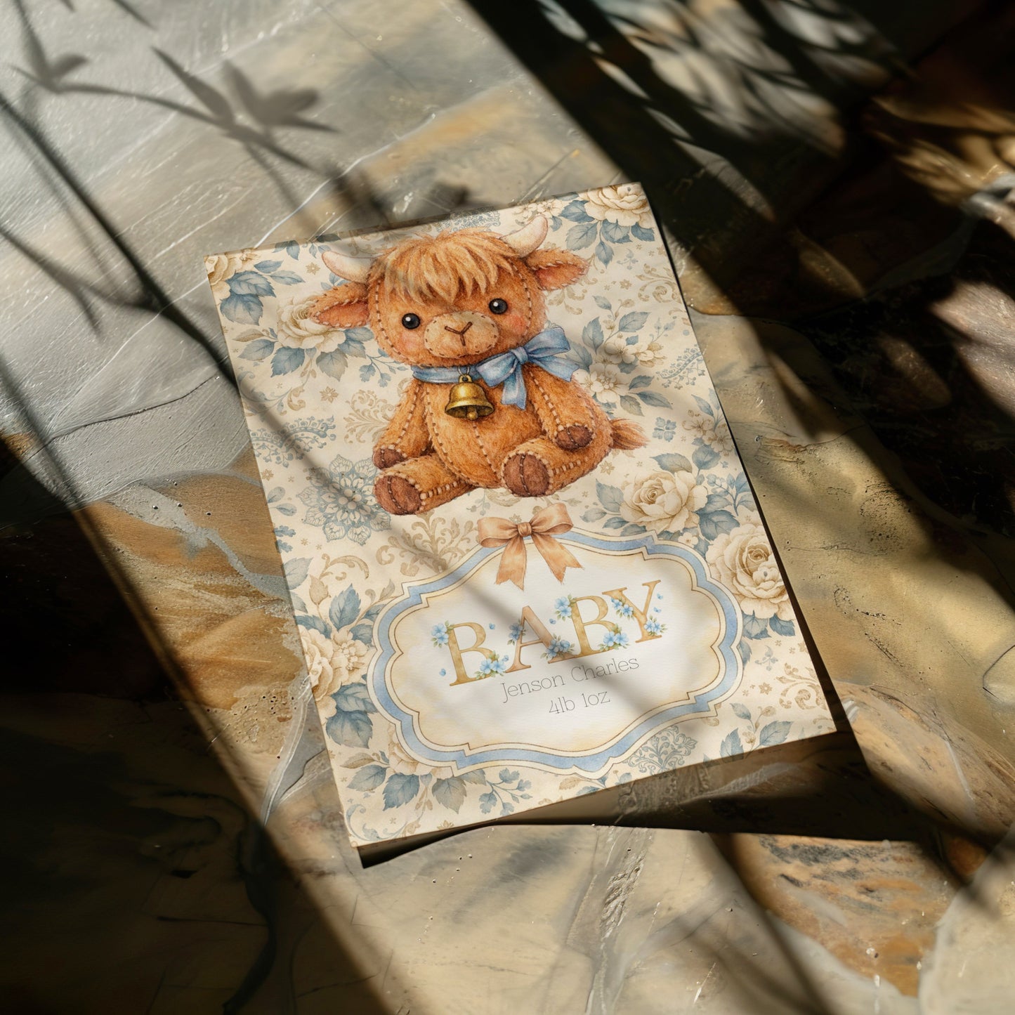 A baby card featuring a cute highland cow with a blue bow and gold bell, set against a floral cream background with the word 'BABY' in gold colouring. The card is photographed on a marble surface with dappled sunlight and shadows.