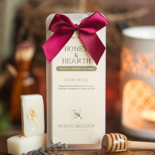 A boxed honey and earth scented wax melt by Robin's Whisper, tied with a magenta satin ribbon, displayed on a wooden surface with a candle, wax melt, and honey dipper in a warm, cosy setting.