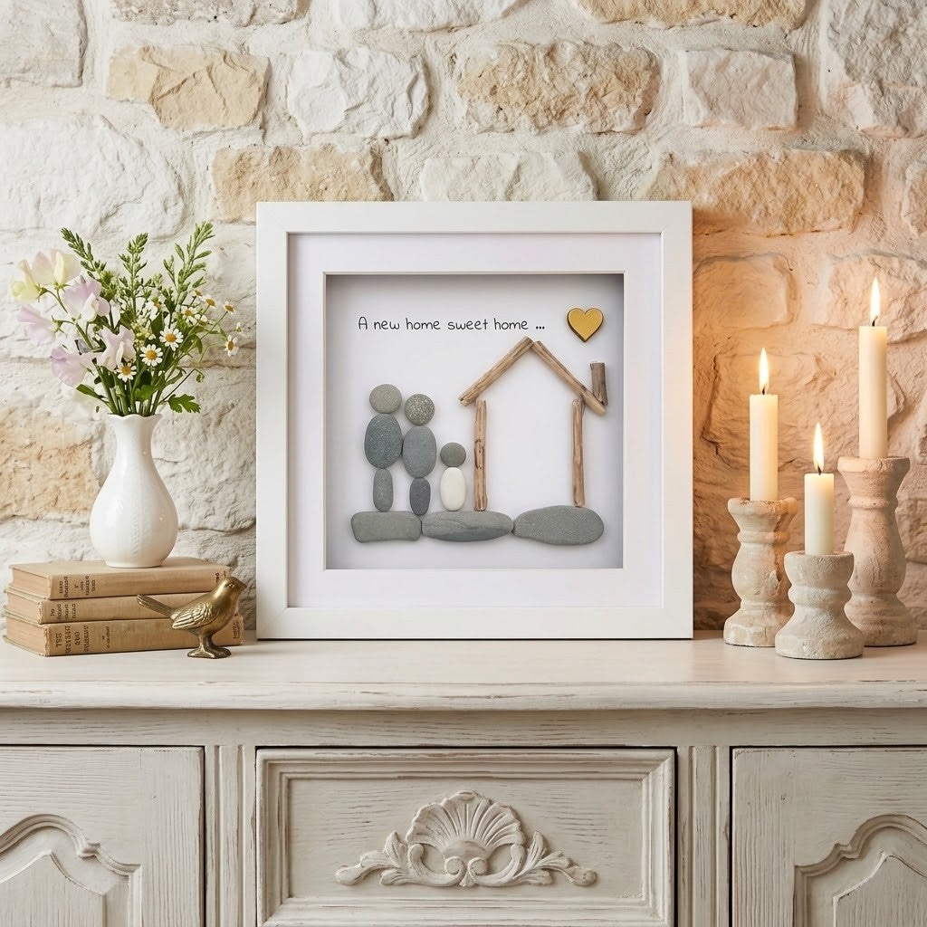 A framed home-themed wall art piece displayed on a light wood cabinet against a stone wall. The white-framed print features a small wooden house, smooth grey stones, a gold heart, and the phrase 'a new home sweet home.