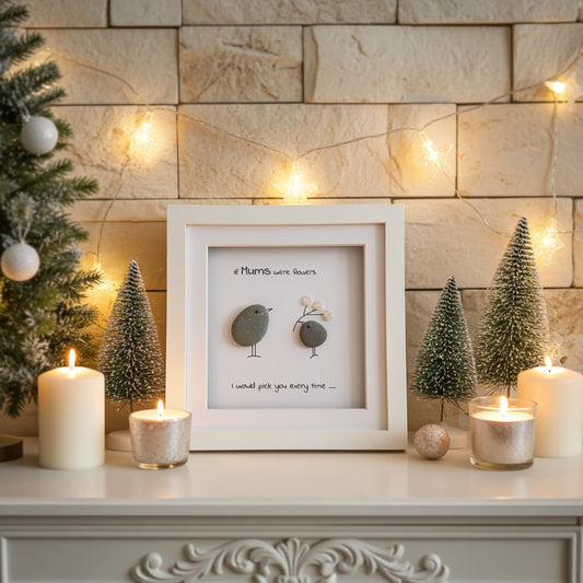Framed pebble artwork with text and small birds on a decorative surface with candles and Christmas trees.