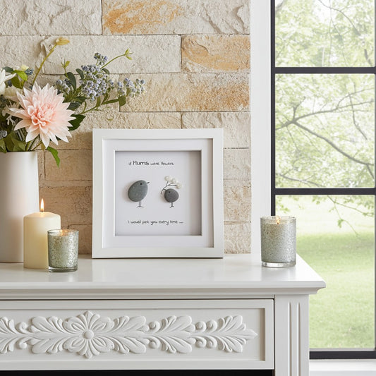 Decorative scene with a framed pebble art picture, candles, and flowers on a white surface.