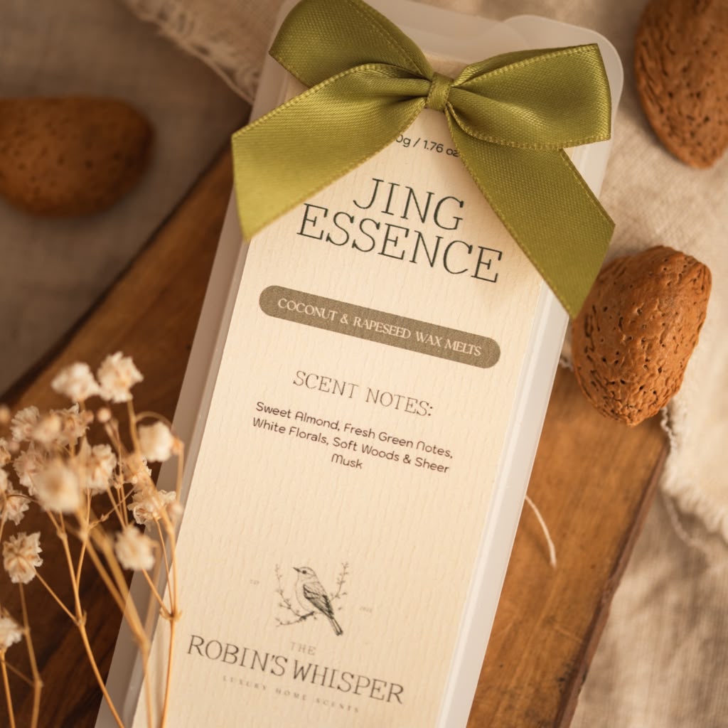 A boxed wax melt product labelled 'JING ESSENCE' by Robin's Whisper, wrapped in a cream paper sleeve with a large olive green ribbon bow, photographed on a wooden surface with dried flowers and textured props around it.