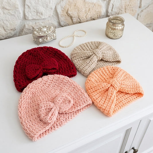 Four knitted baby hats in red, beige, light pink, and peach, arranged on a white table with a stone wall background and decorative props.