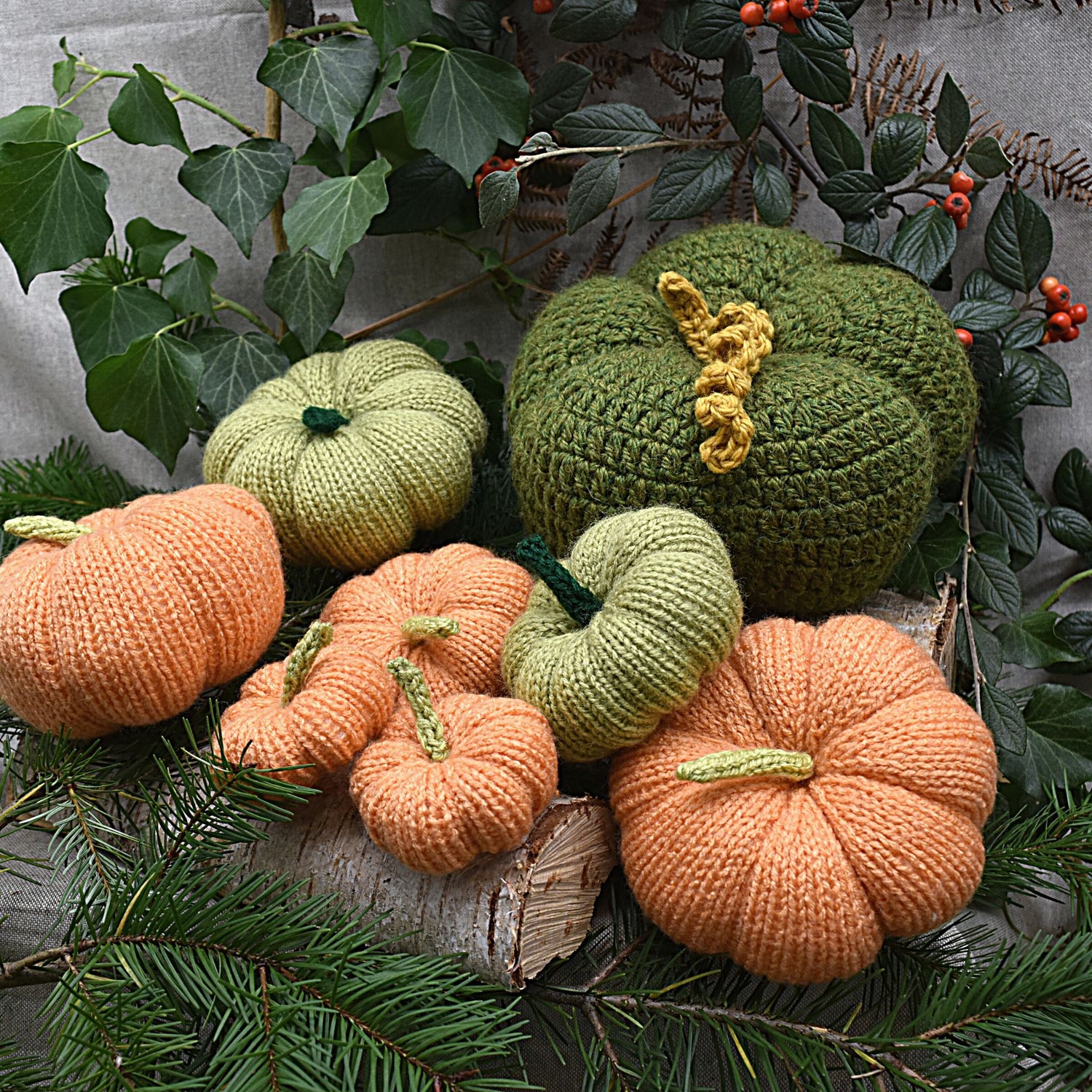 Knitted pumpkin decorations in orange, green, and yellow, displayed with autumnal background and decorative pumpkin-shaped wooden base.