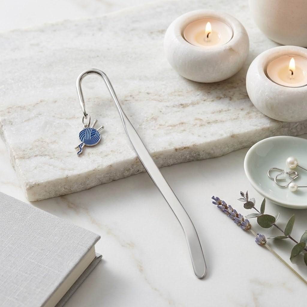 A silver metal bookmark shaped like knitting needles with a small blue and white yarn ball charm, displayed on a white marble slab with lit candles, a book, and small jewelry pieces.