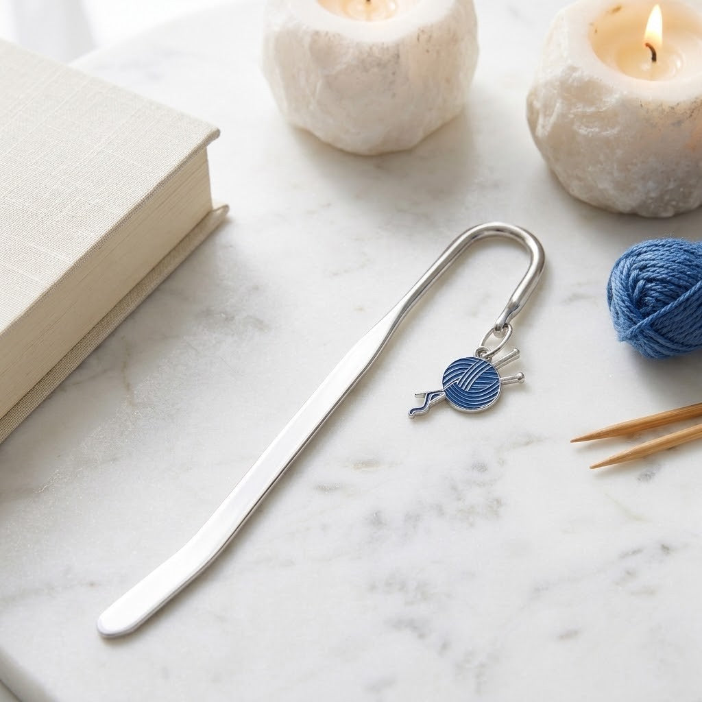 A silver metal bookmark shaped like knitting needles and a blue yarn ball charm, resting on a white marble surface next to a book, candles, and blue yarn.