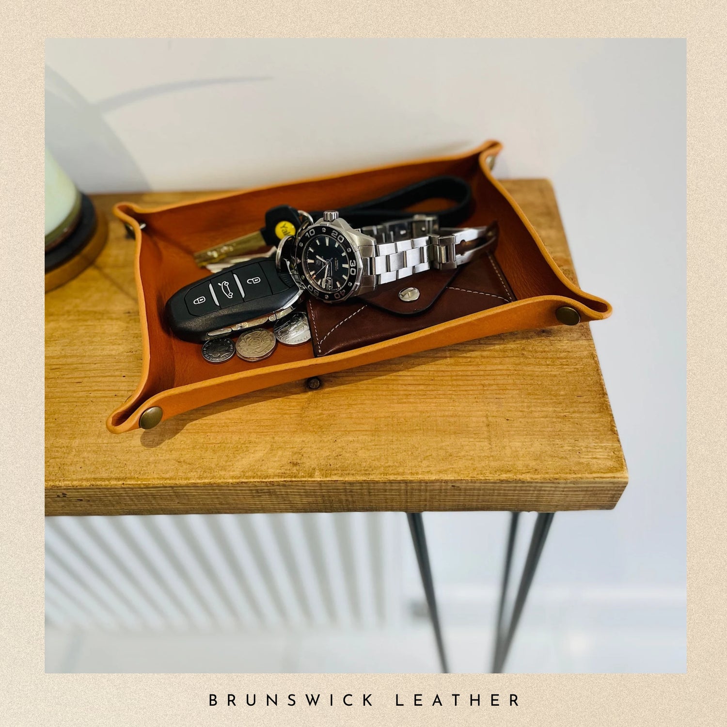 Brown leather tray with keys, coins, and a watch on a wooden surface, branded 'Brunswick Leather'.