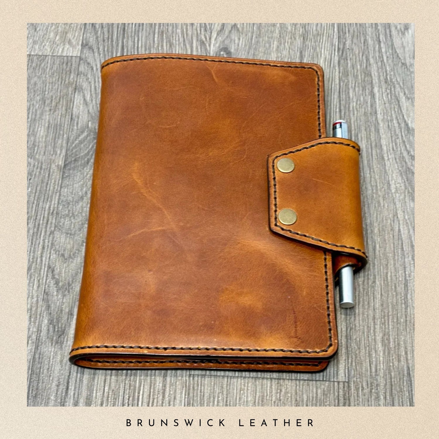 Brown leather notebook with pen holder on a wooden surface, branded 'Brunswick Leather'.