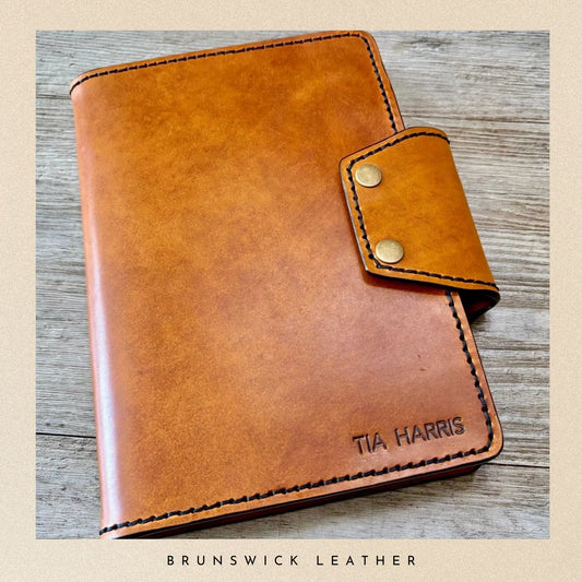 Brown leather notebook with gold accents on a wooden surface, branded 'Tia Harris' and 'Brunswick Leather'.