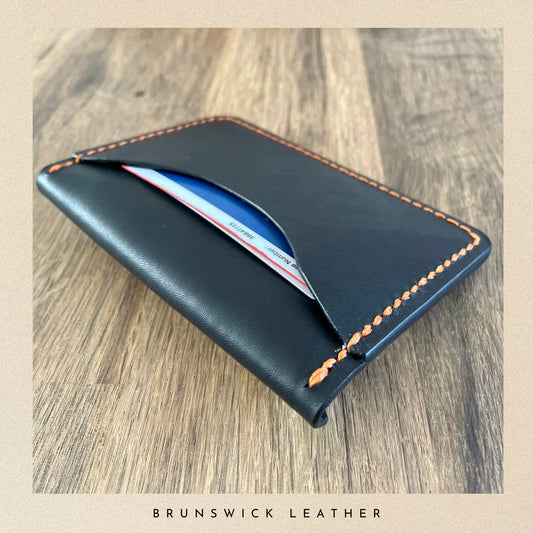 Black leather wallet with orange stitching on a wooden surface, branded 'Brunswick Leather'.