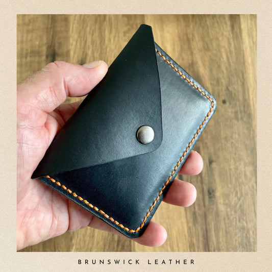 Hand holding a black leather wallet with orange stitching on a wooden surface, branded 'Brunswick Leather'.