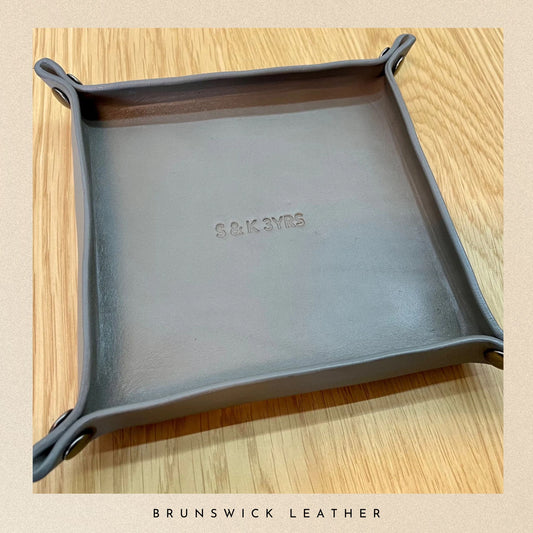 Gray leather tray with 'S&K SYMS' branding on a wooden surface, featuring Brunswick Leather.