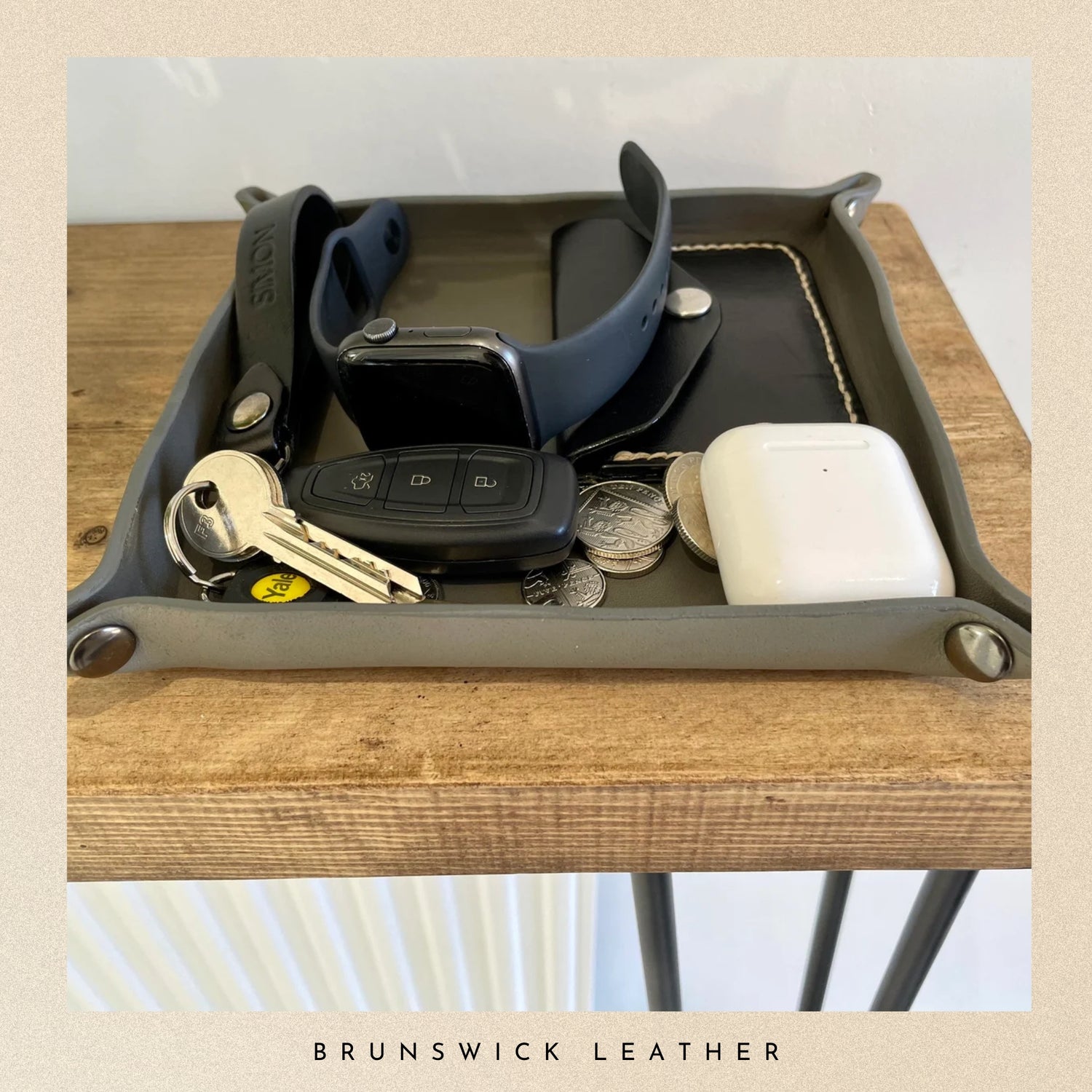 Tray with various items including keys, a car remote, coins, and a phone case on a wooden surface. Brand 'Brunswick Leather' visible.