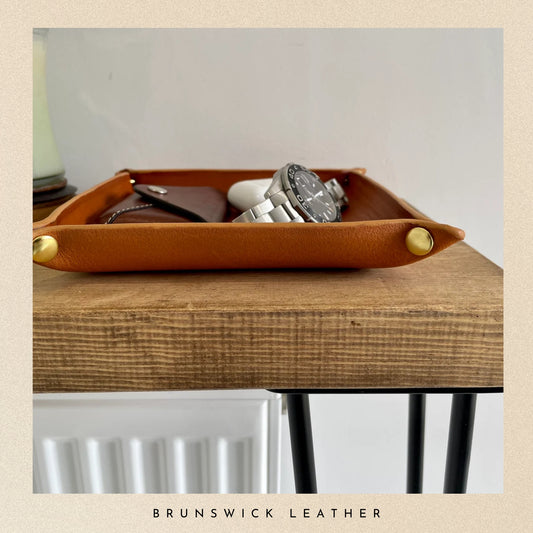 Brown leather tray with a watch and wallet on a wooden surface, branded 'Brunswick Leather'.