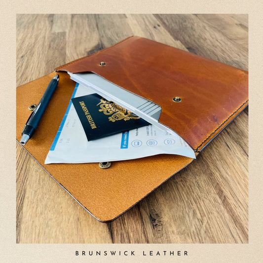 Brown leather wallet with passport, tickets, and pen on a wooden surface, branded 'Brunswick Leather'.