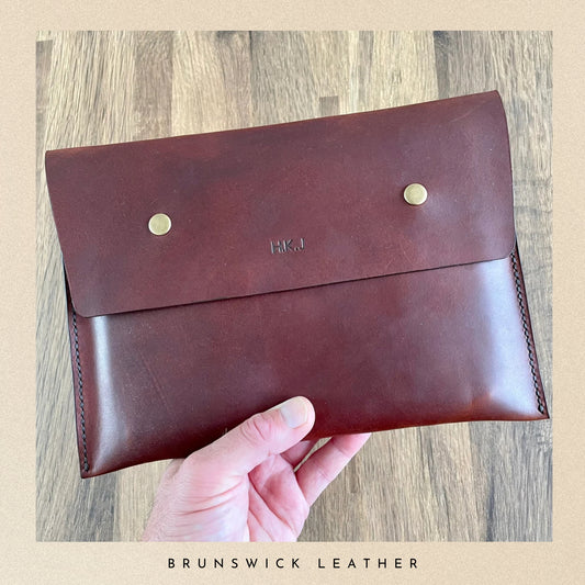 Brown leather clutch with gold buttons held by a hand on a wooden surface, branded 'Brunswick Leather'.