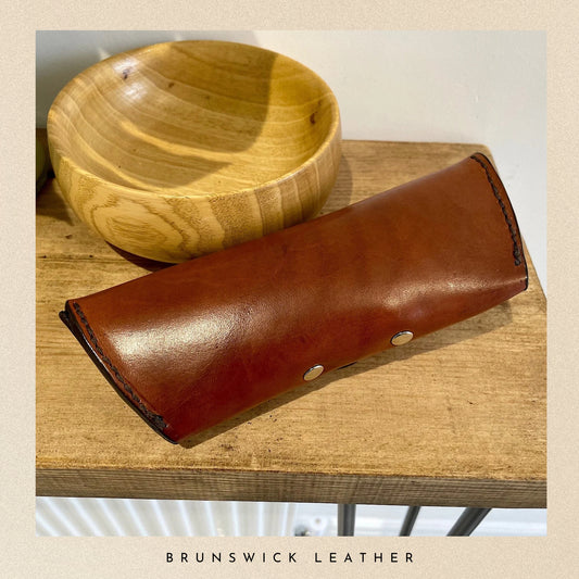 Brown leather case with gold studs on a wooden surface, branded 'Brunswick Leather'.