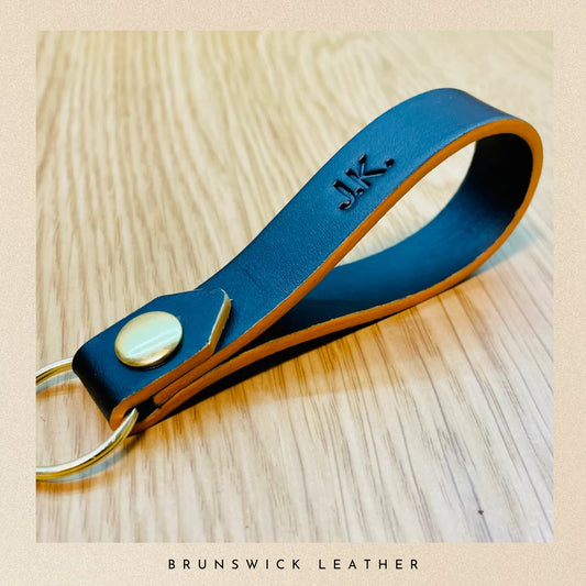 Black leather keychain with gold accent on a wooden surface, branded 'Brunswick Leather'.