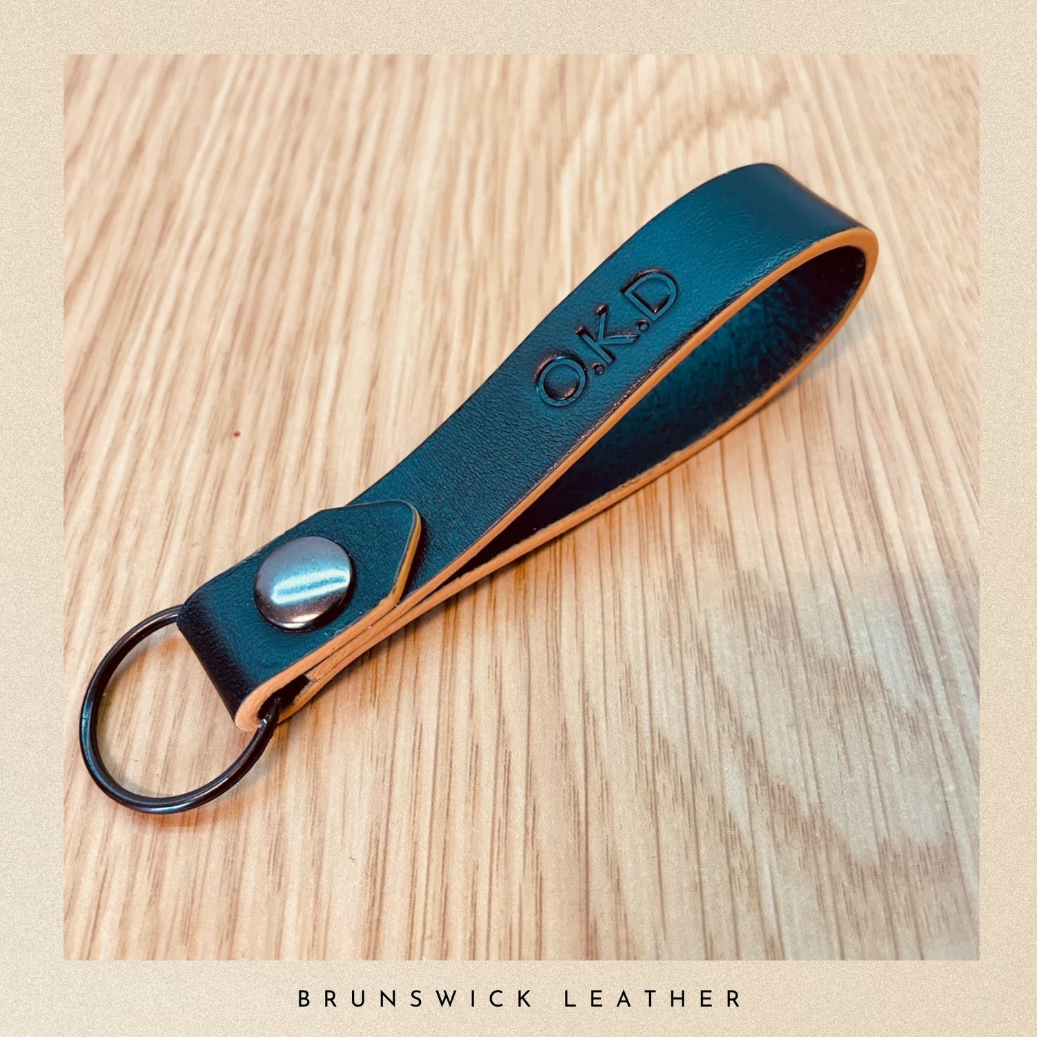 Black leather keychain with 'O.K.D.' engraving on a wooden surface, branded 'Brunswick Leather'.