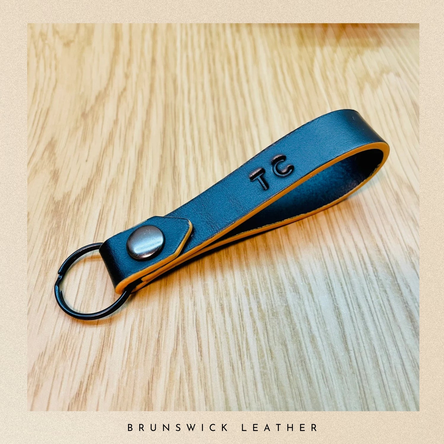 Black leather keychain with initials on a wooden surface, branded 'Brunswick Leather'.
