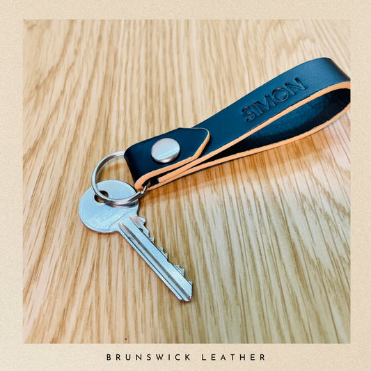 Keychain with a black leather strap and silver key on a wooden surface, branded 'Brunswick Leather'.
