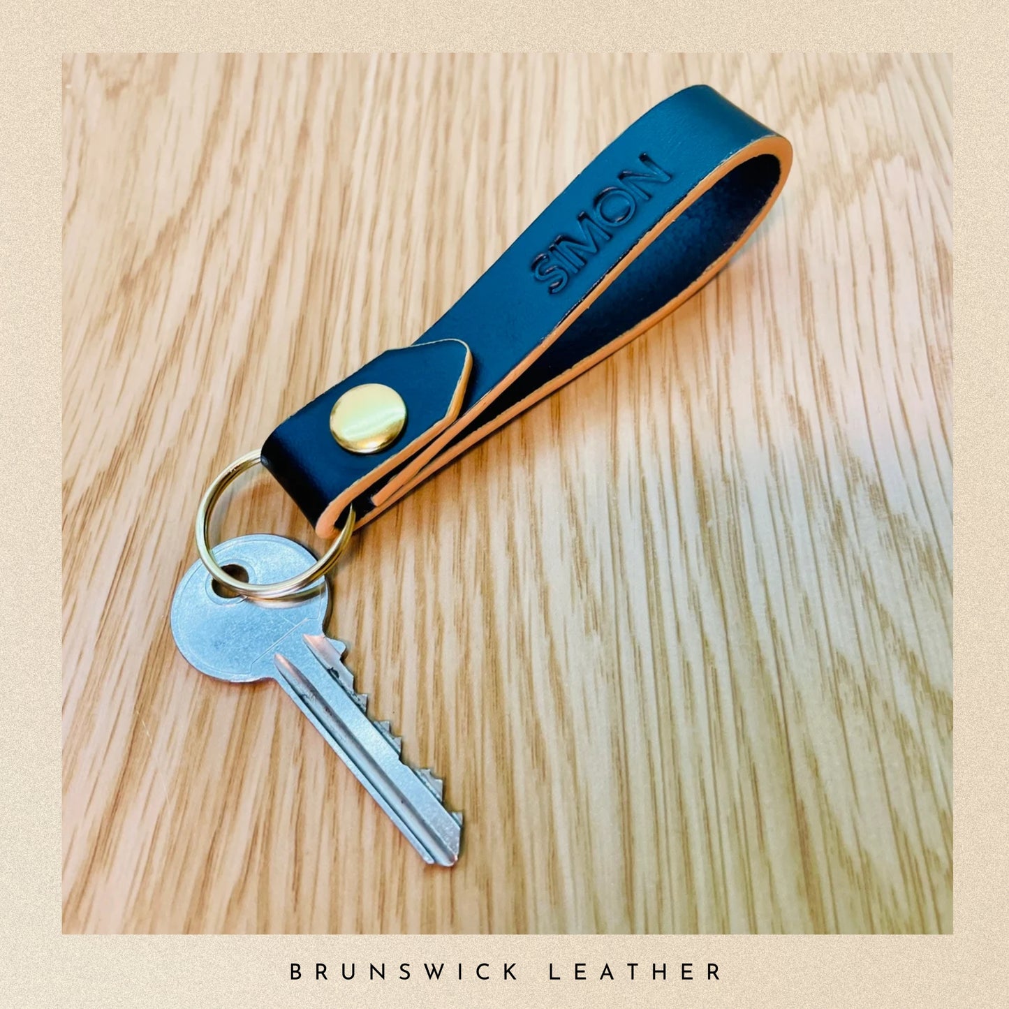 Black leather keychain with a key on a wooden surface, branded 'Brunswick Leather'.