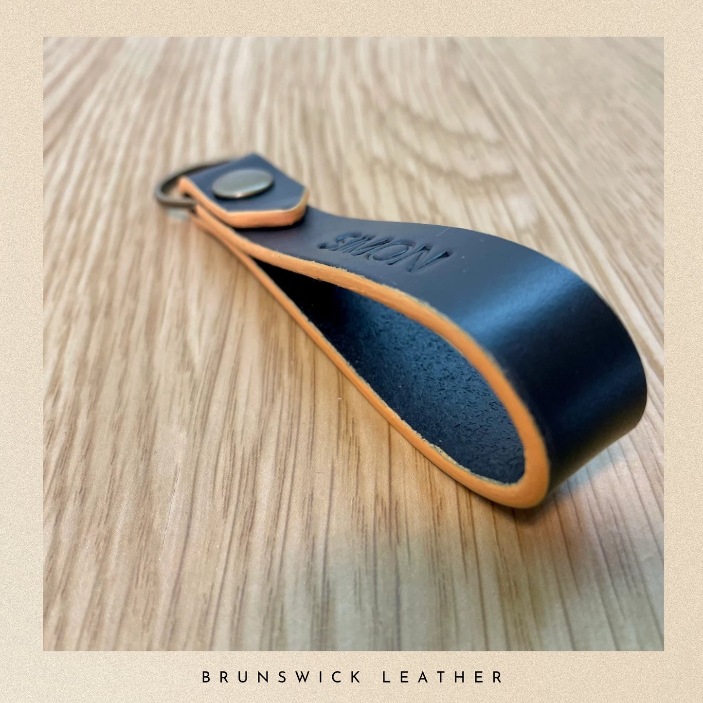 Black leather keychain with orange trim on a wooden surface, branded 'Brunswick Leather'.