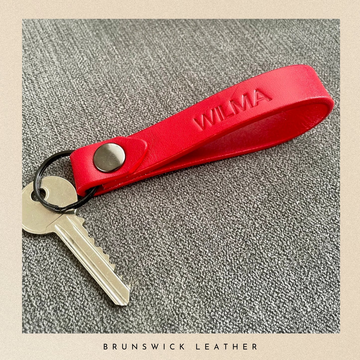 Red leather keychain with a key on a textured surface, branded 'Wilma' and 'Brunswick Leather'.