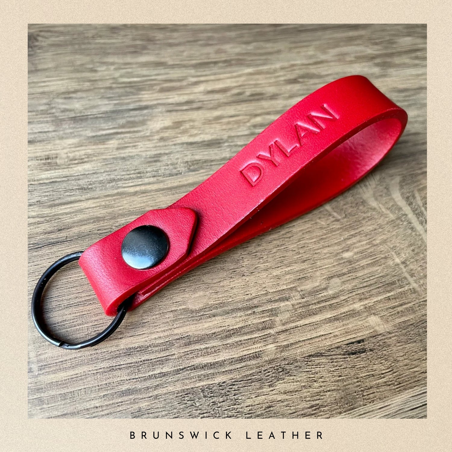 Red leather keychain with 'DYLAN' engraving on a wooden surface, branded 'Brunswick Leather'.